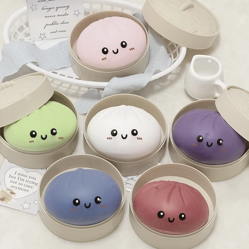 Six squishys bao mats posés dans leurs paniers vapeur en bambou, teintes rose, vert, blanc, violet, bleu et rouge, sur un fond sable.